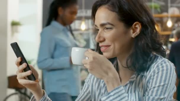 美しい女性がカフェを使用してスマート フォン 彼女の笑顔で座っています 彼女のテーブル カップ飲料とクロワッサン バック グラウンドで忙しいスタイリッシュなコーヒー ハウス — ストック動画