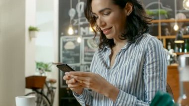 Güzel kadın Cafe kullanır Smartphone içinde oturur. Her arkadaş mesajlaşma ya da sevilen bir süre gülümsüyor. İçecek ve kruvasan ile onun tablo bardakta. Arka planda şık kahve evi meşgul.