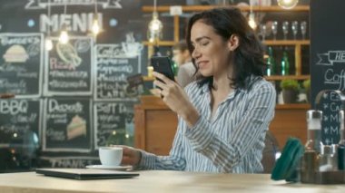 Güzel kadın Cafe kullanır Smartphone içinde oturur. Her arkadaş mesajlaşma ya da sevilen bir süre gülümsüyor. İçecek bir bardaktan içiyor. Arka planda şık kahve evi meşgul.