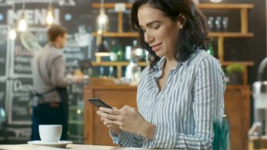 Güzel kadın Cafe kullanır Smartphone içinde oturur. Her arkadaş mesajlaşma ya da sevilen bir süre gülümsüyor. İçecek ile onun tablo bardakta. Arka planda şık kahve evi meşgul.