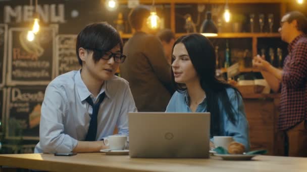 英俊的亚洲男人和美丽的白种人年轻女子坐在咖啡厅工作在一台笔记本电脑上 在后台其他客户在时尚的环境中 — 图库视频影像