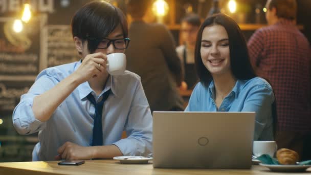 英俊的亚洲男人和美丽的白种人年轻女子坐在咖啡厅工作在一台笔记本电脑上 在后台其他客户在时尚的环境中 — 图库视频影像