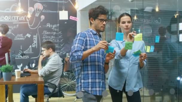 Kreative männliche und weibliche Entwickler nutzen Glaswände, um neue Projekte mit klebrigen Papieren zu skizzieren. Im Hintergrund talentierte junge Leute, die mit der Tafel und am PC arbeiten.