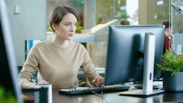 彼女のデスク パソコン上の作品に美しい若い白人女性の座っています 同僚の作業と背景の忙しいオフィスで — ストック動画