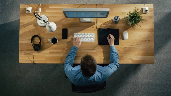 顶视图的一个年轻的平面设计师 工作在数字绘图平板电脑上坐在他的桌子 另外还有桌面计算机 耳机和智能手机在桌子上 — 图库照片