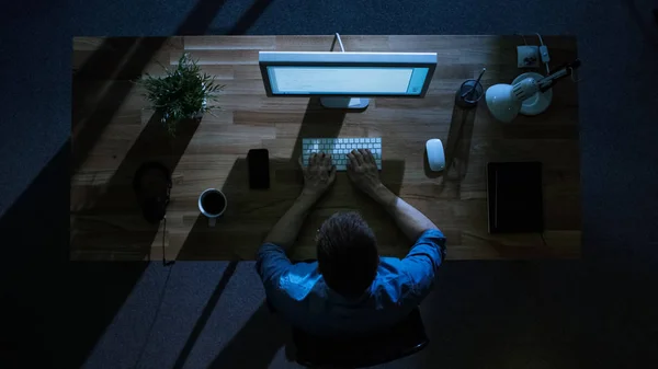 Top View of Male Programmer Working at His Desktop Computer at Night. His Table is Illuminated by Cold Blue Light From Outside.