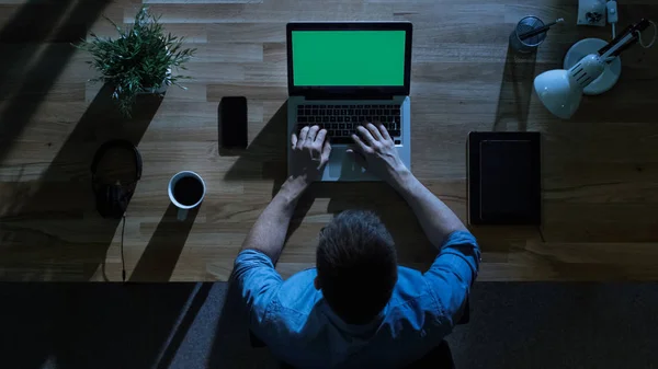 Top View of Male Student Working at His Desktop Computer with Mock-up Green Screen at Night. His Table is Illuminated by Cold Blue Light From Outside.
