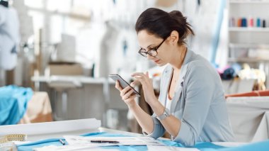 Güzel kadın moda, tasarımcı, terzilik manken, bluz, ayarlama kullanma Smartphone ile çalışma. Onun stüdyom Bright ve Sunny, giysi asılı, renkli kumaşlar yalan masada.