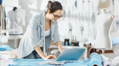 Güzel kadın Tasarımcısı kullanır Laptop onun masasında, çeşitli kumaşlar ve dikiş öğeleri ile kaplıdır. Onun Studio Sunny, manken, Giysi asılı ve skeçler Pinned duvara.