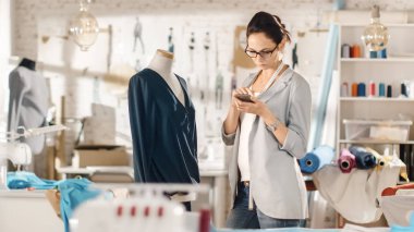 Güzel kadın moda, tasarımcı, terzilik manken, bluz, ayarlama kullanma Smartphone ile çalışma. Onun stüdyom Bright ve Sunny, giysi asılı, renkli kumaşlar yalan masada.