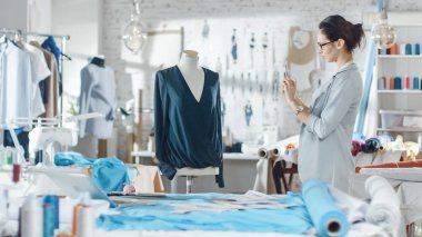Güzel kadın moda, tasarımcı, terzilik manken, bluz, ayarlama kullanma Smartphone ile çalışma. Onun stüdyom Bright ve Sunny, giysi asılı, renkli kumaşlar yalan masada.