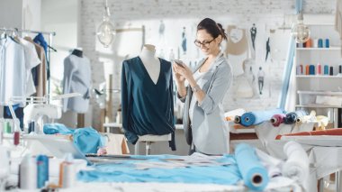Güzel kadın moda, tasarımcı, terzilik manken, bluz, ayarlama kullanma Smartphone ile çalışma. Onun stüdyom Bright ve Sunny, giysi asılı, renkli kumaşlar yalan masada.