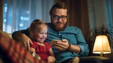 Akşam baba ve küçük kız oturan bir kanepe oturma odası seyretmek smartphone cep telefonu ile komik videolar.