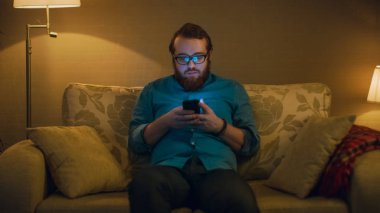 Bir adam onun oturma odasında kanepede oturmuş, Smartphone kullanarak portre vurdu. Zemin lambaları döndü yanıyor.