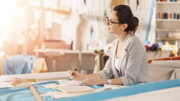 Beautiful Female fashion, designer,  Sits at Her Desk and Draws Sketches. Her Studio is Sunny, Full of Colorful Details, Fabrics, Sewing Items, Sketches.