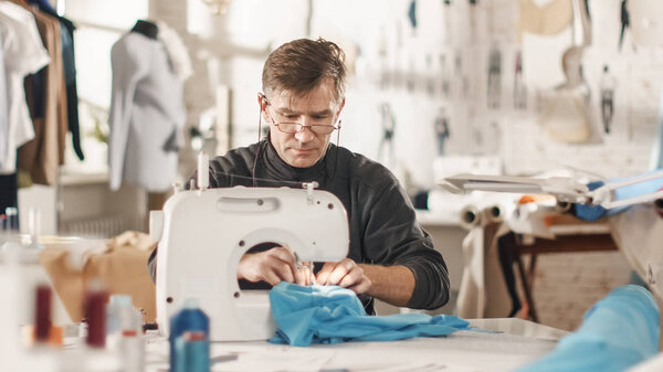 Male fashion, designer, / Dressmaker Working on a Sewing Machine in His Sunny Studio. Various Sewing items and Fabrics Laying Around Him, Visible Sketches Pinned to the Wall.