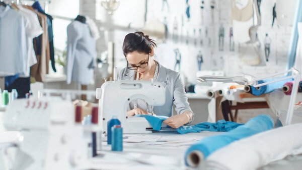 Female fashion, designer, / Seamstress/ Dressmaker Working on a Sewing Machine in Her Sunny Studio. Various Sewing items and Fabrics Laying Around Her, Visible Sketches Pinned to the Wall.