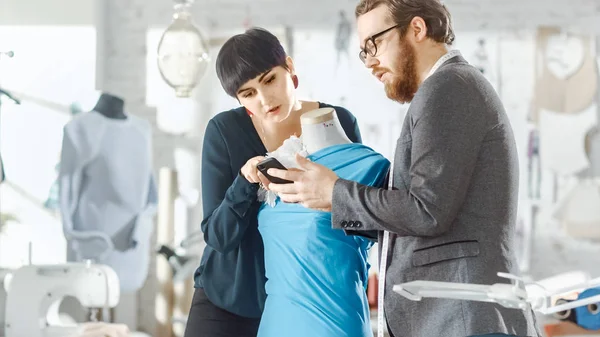 Male and Female fashion, designer, s are Working on Tailored Mannequin Wearing Clothes. They Consult Smartphone. In Their Studio Sewing Machine, Fabrics and Sketches are Visible.