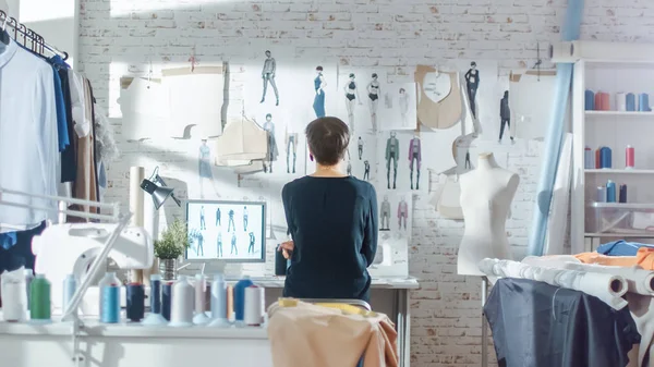 Female fashion, designer,  Looking at Drawings and Sketches that are Pinned to the Wall. Studio is Sunny. Colorful Fabrics, Clothes Hanging and Sewing Items are Visible.