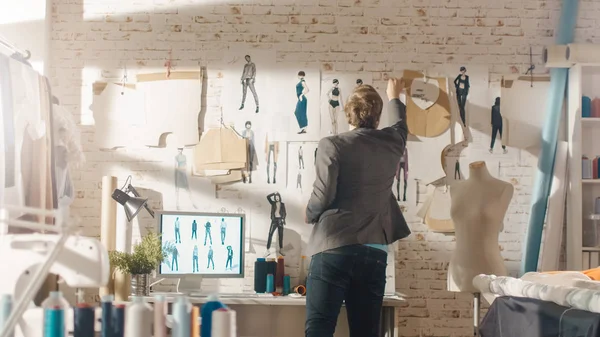 男时尚 设计师 把衣服的素描钉在墙上 他的工作室是晴天 个人电脑显示他的工作 彩色布料 挂衣和缝纫物品可见 — 图库照片