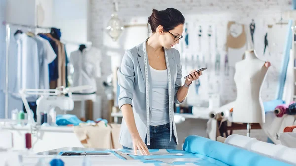 Güzel esmer moda, tasarımcı, kumaşlar ve Her Smartphone danışır şablonları bırakır. O bir ışık renkli stüdyoda çalışıyor tam çeşitli giysiler, kumaşlar ve duvardaki çizimler.