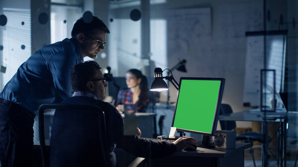 Late at Night Two Men Discuss about Green Screen Template that are Shown on Display of a Personal Computer. Office Looks Modern with Lots of Blueprints on the Walls. In the Background Woman Working.