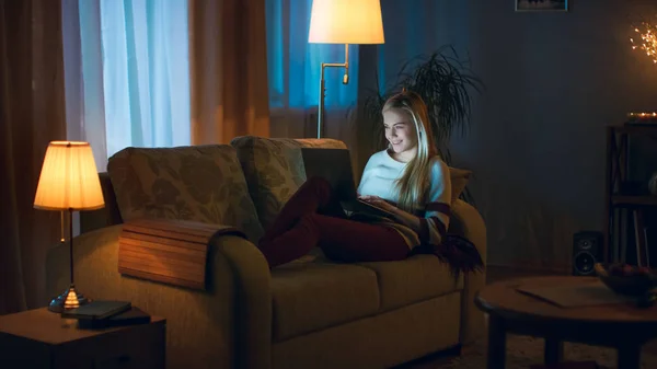 夕方には美しい若い女性は 彼女の膝の上ノート パソコンとソファの上あります 彼女はタイプする 楽しい 暖かく 居心地の良い部屋に見える — ストック写真