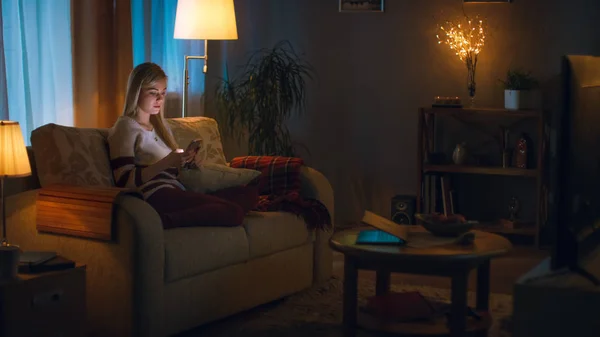 In the Evening Cute Little Girl is Sitting in Living Room on a Sofa, She Watches Television. She Holds Her Soft Toy, Room Lights are On.