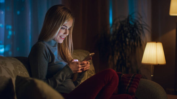 Beautiful Young Woman Sitting on Her Sofa in a Living Room. She Uses Smartphone with Interest. It's Evening, Big City is Seen in the Window.