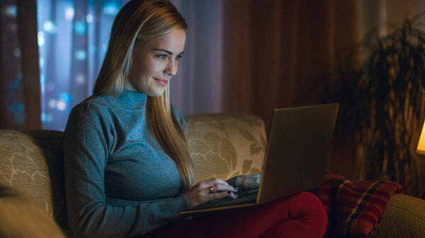 Beautiful Young Woman Works on a Laptop in Her Living Room, She is Sitting on a Sofa with Notebook on Her Lap. In the Window Big City is Seen.