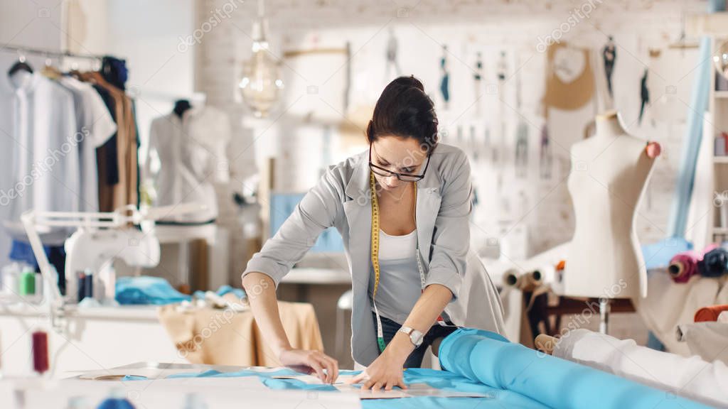 Beautiful Brunette fashion, designer,  Lays Out Templates on the fabrics and Looks at Her Smartphone. She Works in a Light Colorful Studio Full of Various Clothes, Fabrics and Sketches on the Wall.