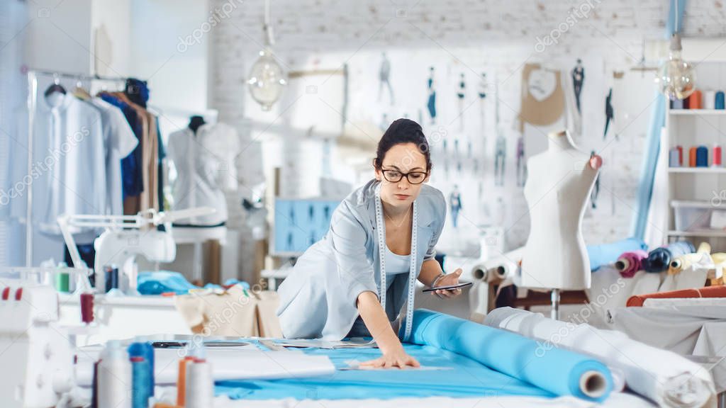 Beautiful Brunette fashion, designer,  Lays Out Templates on the fabrics and Consults Her Smartphone. She Works in a Light Colorful Studio Full of Various Clothes, Fabrics and Sketches on the Wall.