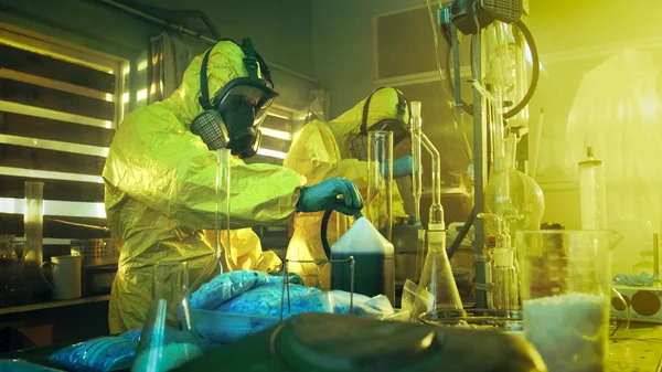 在地下实验室两位身穿防护服和面具的秘密化学家煮药物 他们工作与烧杯 蒸馏玻璃器皿 滤毒罐和軟 真实犯罪概念 — 图库照片