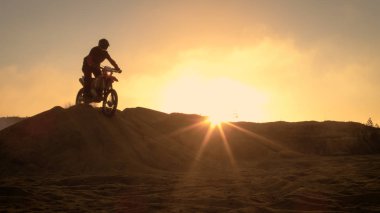 Professional Motocross Motorcycle Rider Drives Over the Dune and Further Down the Off-Road Track. It's Sunset and Track is Covered with Smoke/ Mist. clipart