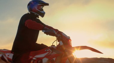 Professional FMX Motorcycle Rider Prepares to Start Driving on His Bike Over Hard Sandy Off-Road Terrain. Sun is Setting. clipart