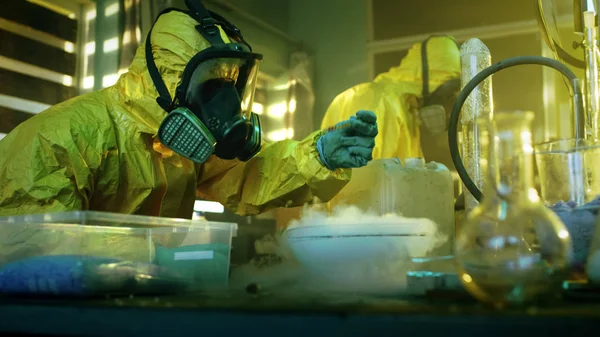 地下薬物研究室で つの秘蜜化学防護マスクとカバーオールを着て調理された薬剤の純度と強度をテストします 彼らは放棄で働くガラスの建物と調理器具 — ストック写真