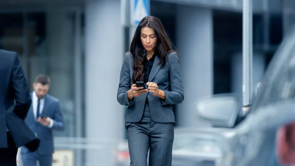 商务女性穿着量身定做的西装 在商业区繁华的大城市街道上行走 检查她的智能手机 自信的女人在她的方式做大生意 — 图库照片