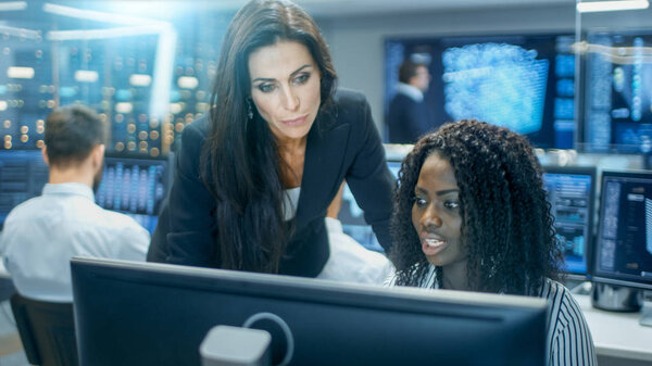 Female Team Leader Consults Young Computer Engineer. They Work in a Crowded Office on a Neural Network/ Artificial Intelligence Project.