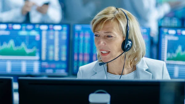 Chief Sales Force Representative Talks into the Headset. Behind Her People Working, Screens Show Stock Market Ticker Numbers and Graphs.