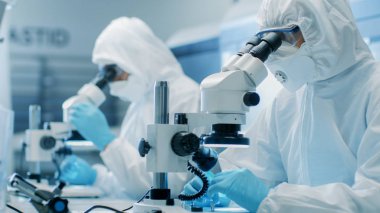 İki mühendis / bilim adamları / teknisyenleri steril temiz oda takım elbiseli kullanın mikroskoplar bileşen ayarı ve araştırma için. Onlar bir elektron komponentler fabrika üretim iş.