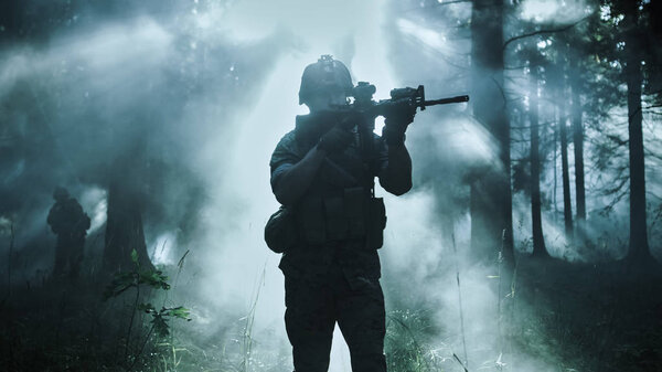 Silhouette of the Fully Equipped Soldier Moving Through Smokey Forest with Rifle Ready To Shoot. Reconnaissance Military Operation. Squad Moving Behind Him. Dark and Cold Ambient.