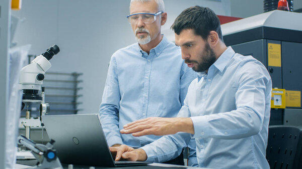 Two Industrial Scientists/ Engineers/ Technologists Have Discussion while Working on Laptop Computer. In the Background Modern Laboratory/ Factory Equipment.