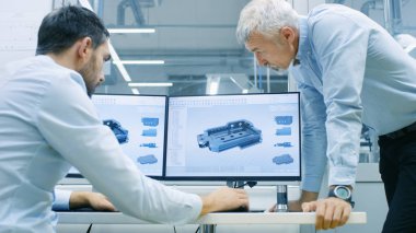 Endüstriyel tasarımcı konuşma Kıdemli mühendis ile yeni bileşen tasarımı Cad programında çalışırken vardır. O inşaat üstünde kişisel bilgisayar ile iki monitör.
