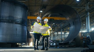 Male and Female Industrial Engineers Look at Project Blueprints While Standing Surround By Pipeline Parts in the Middle of Enormous Heavy Industry Manufacturing Factory clipart