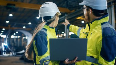 İçinde ağır endüstriyel fabrika kadın mühendis Laptop tutar ve tartışma ile proje yöneticisi vardır. Sabit şapka ve emanet ceketler giyiyorlar. Arka plan kaynak / devam eden metal işi.