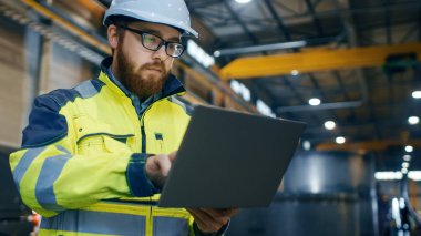 Industrial Engineer in Hard Hat Wearing Safety Jacket Uses Touchscreen Laptop. He Works at the Heavy Industry Manufacturing Factory.