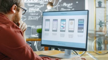 Erkek mobil oyun uygulama geliştirici tel kafes tasarımı bir kişisel bilgisayarda çalışır. Onun iş arkadaşı proje planı yaratıcı Office Blackboard duvara çizer..