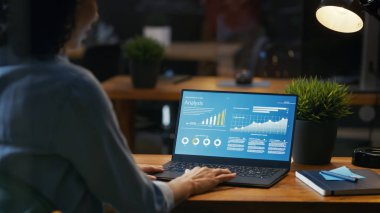 Onun masasında kadın analisti istatistikleri, grafikler ve gösterilen bir dizüstü bilgisayarda çalışır. O yaratıcı ofiste ahşap tabloda çalışıyor. Omuz görüntüleri.