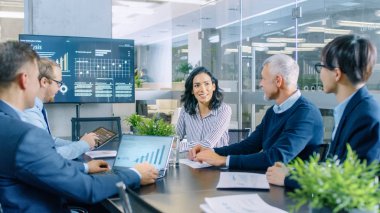 Kadın CEO toplantı odasında iş arkadaşları için bir rapor verir. Çeşitli akıllı kişilik ekibin dikkatle dinler. Modern arıyorsunuz ofis duvar istatistikleri gösterilen Tv var.