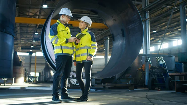 Male and Female Industrial Engineers in Hard Hats Discuss New Project while Using Tablet Computer. They\'re Making Calculated Engineering Decisions.They Work at the Heavy Industry Manufacturing Factory.